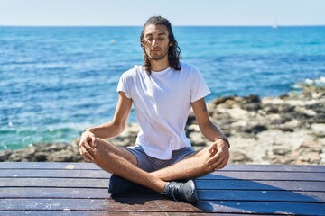 Young hispanic man doing yoga exercise sitting on bench at seaside