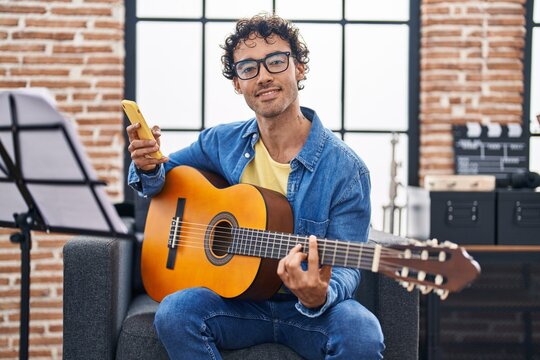 Young hispanic man musician playing classical guitar using smartphone at music studio