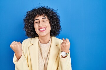 Obraz na płótnie Canvas Young brunette woman with curly hair standing over blue background very happy and excited doing winner gesture with arms raised, smiling and screaming for success. celebration concept.