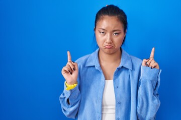 Asian young woman standing over blue background pointing up looking sad and upset, indicating direction with fingers, unhappy and depressed.