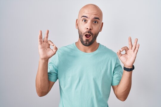 Middle age bald man standing over white background looking surprised and shocked doing ok approval symbol with fingers. crazy expression