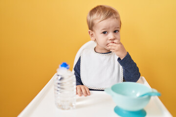 Adorable blond toddler sitting on highchair eating snack over isolated yellow background