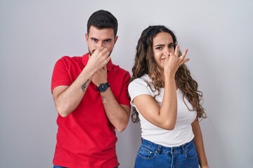 Young hispanic couple standing over isolated background smelling something stinky and disgusting, intolerable smell, holding breath with fingers on nose. bad smell - obrazy, fototapety, plakaty