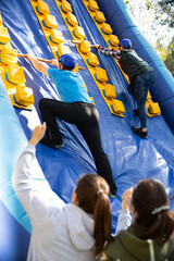 Group of enthusiastic men and women passing obstacles and having fun on inflatable arena at...