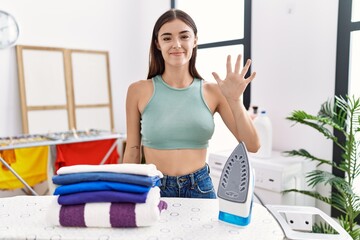 Young hispanic woman ironing clothes at laundry room showing and pointing up with fingers number five while smiling confident and happy.