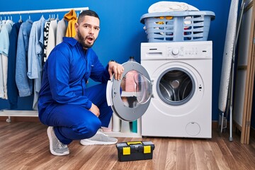 Hispanic repairman working on washing machine scared and amazed with open mouth for surprise, disbelief face