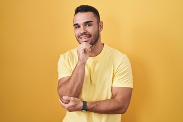 Young hispanic man standing over yellow background with hand on chin thinking about question, pensive expression. smiling and thoughtful face. doubt concept.