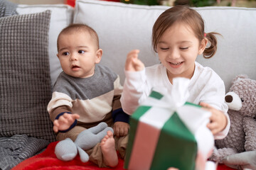 Brother and sister opening gift sitting on sofa by christmas tree at home