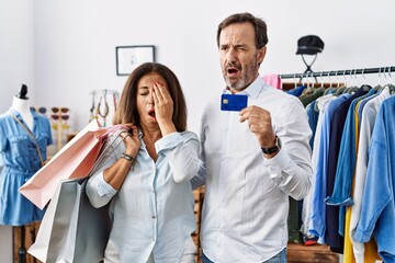 Hispanic middle age couple holding shopping bags and credit card yawning tired covering half face, eye and mouth with hand. face hurts in pain.