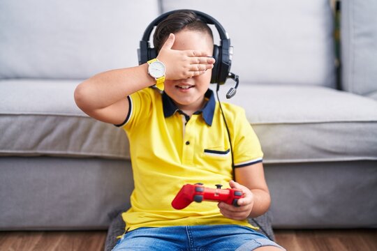 Young Hispanic Kid Playing Video Game Holding Controller Wearing Headphones Smiling And Laughing With Hand On Face Covering Eyes For Surprise. Blind Concept.