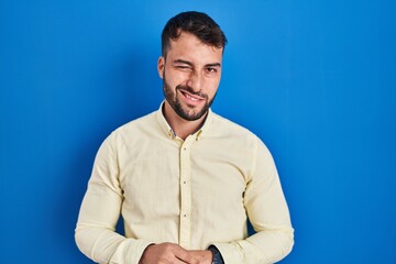 Handsome hispanic man standing over blue background winking looking at the camera with sexy expression, cheerful and happy face.