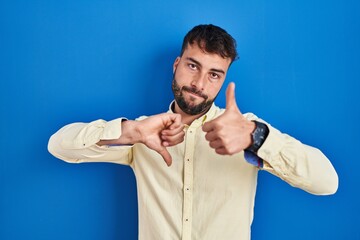 Handsome hispanic man standing over blue background doing thumbs up and down, disagreement and agreement expression. crazy conflict