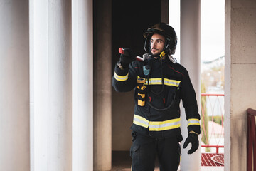 A young and uniformed firefighter walking inside a building that has burned carrying a mace on his...