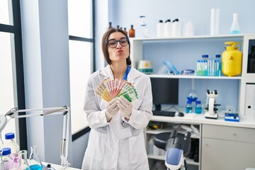 Young hispanic woman working at scientist laboratory holding money banknotes looking at the camera blowing a kiss being lovely and sexy. love expression.