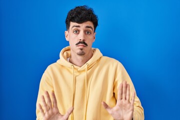Hispanic man standing over blue background moving away hands palms showing refusal and denial with...