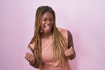 African american woman with braided hair standing over pink background very happy and excited doing winner gesture with arms raised, smiling and screaming for success. celebration concept.