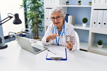 Senior woman with grey hair wearing doctor uniform holding prescription pills smiling happy pointing with hand and finger