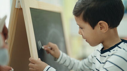 Adorable hispanic boy preschool student writing on blackboard at kindergarten
