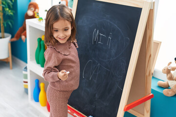 Adorable hispanic girl preschool student smiling confident writing name on blackboard at...