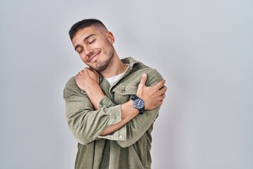 Young hispanic man standing over isolated background hugging oneself happy and positive, smiling confident. self love and self care - obrazy, fototapety, plakaty