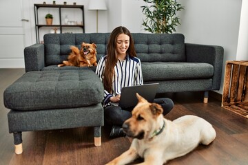 Young hispanic woman using laptop sitting on floor with dogs at home