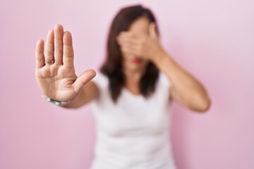 Middle age brunette woman standing over pink background covering eyes with hands and doing stop gesture with sad and fear expression. embarrassed and negative concept.