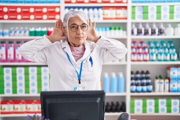 Middle age woman with tattoos working at pharmacy drugstore trying to hear both hands on ear gesture, curious for gossip. hearing problem, deaf