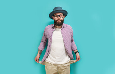 Emotional hipster guy in a hat and streetwear shows empty pockets. The concept of poverty and unemployment. Indoor studio shot against a blue background.