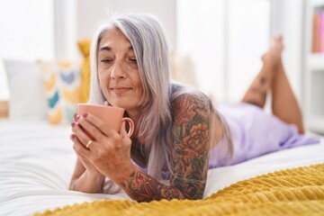 Obraz na płótnie Canvas Middle age grey-haired woman drinking cup of coffee lying on bed at bedroom