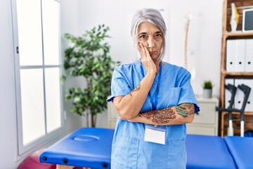 Middle age grey-haired woman wearing physiotherapist uniform at medical clinic thinking looking tired and bored with depression problems with crossed arms.