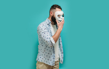 Portrait of a man with a beard covering his face with a white mask hiding his real mysterious identity. Hypocrisy, fraud, lie and insincerity concept. Indoor studio shot against a blue background. - obrazy, fototapety, plakaty