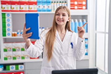 Young caucasian woman working at pharmacy drugstore doing video call with tablet smiling happy pointing with hand and finger to the side
