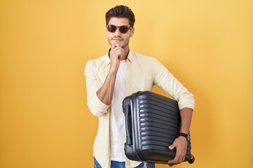 Young hispanic man holding suitcase going on summer vacation with hand on chin thinking about question, pensive expression. smiling and thoughtful face. doubt concept.