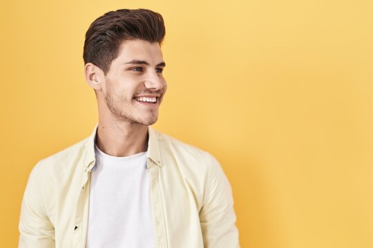 Young hispanic man standing over yellow background looking away to side with smile on face, natural expression. laughing confident.