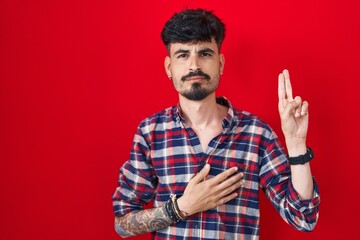 Fototapeta premium Young hispanic man with beard standing over red background smiling swearing with hand on chest and fingers up, making a loyalty promise oath