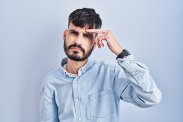 Young hispanic man with beard standing over blue background pointing unhappy to pimple on forehead, ugly infection of blackhead. acne and skin problem