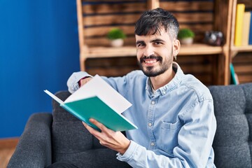 Young hispanic man reading book sitting on sofa at home