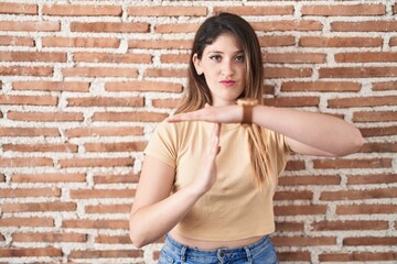Young brunette woman standing over bricks wall doing time out gesture with hands, frustrated and...