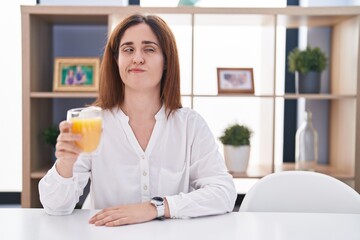 Obraz na płótnie Canvas Brunette woman drinking glass of orange juice smiling looking to the side and staring away thinking.