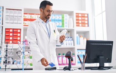 Young hispanic man pharmacist holding pills using credit card and dataphone at pharmacy