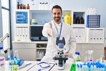 Young hispanic man with beard working at scientist laboratory smiling friendly offering handshake as greeting and welcoming. successful business.