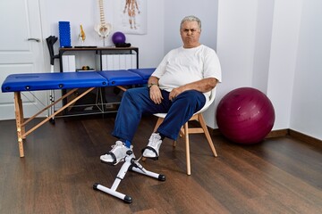 Senior caucasian man at physiotherapy clinic using pedal exerciser puffing cheeks with funny face. mouth inflated with air, crazy expression.