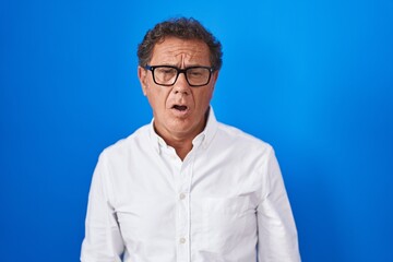 Middle age hispanic man standing over blue background afraid and shocked with surprise and amazed expression, fear and excited face.