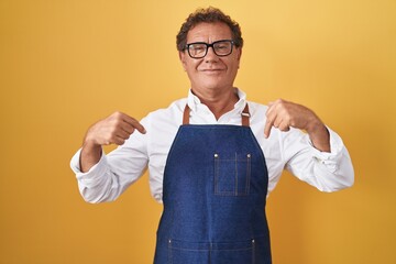 Middle age hispanic man wearing professional cook apron looking confident with smile on face, pointing oneself with fingers proud and happy.
