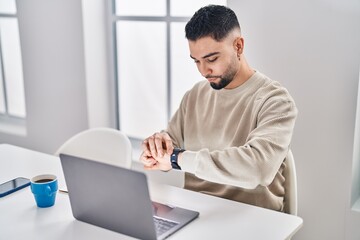 Young arab man looking watch sitting on table working at home