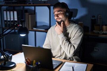 Young handsome man working using computer laptop at night looking confident at the camera smiling with crossed arms and hand raised on chin. thinking positive.