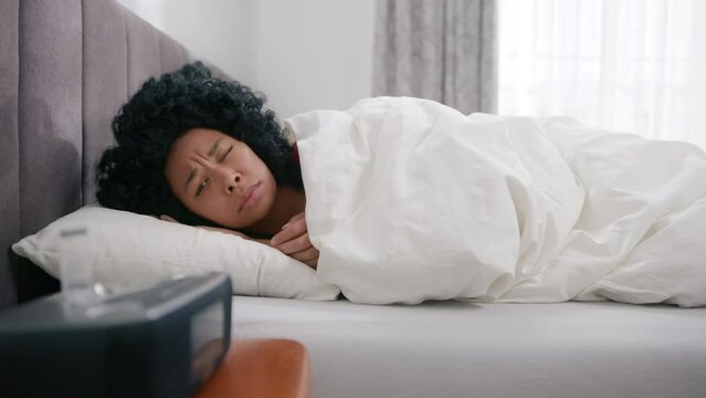 Young african american female sleeping in bed wakes up early hour to disable activated digital alarm clock sounding on bedside table. Black girl covers up in blanket and continue to sleep slow motion