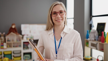 Young blonde woman preschool teacher smiling confident holding books at kindergarten