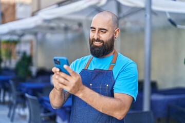 Young bald man waiter smiling confident using smartphone at coffee shop terrace