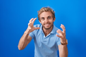 Caucasian man standing over blue background shouting frustrated with rage, hands trying to strangle, yelling mad